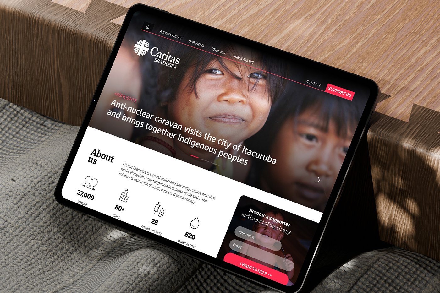 The Caritas Brasileira homepage open on a tablet resting on a sofa, in afternoon light