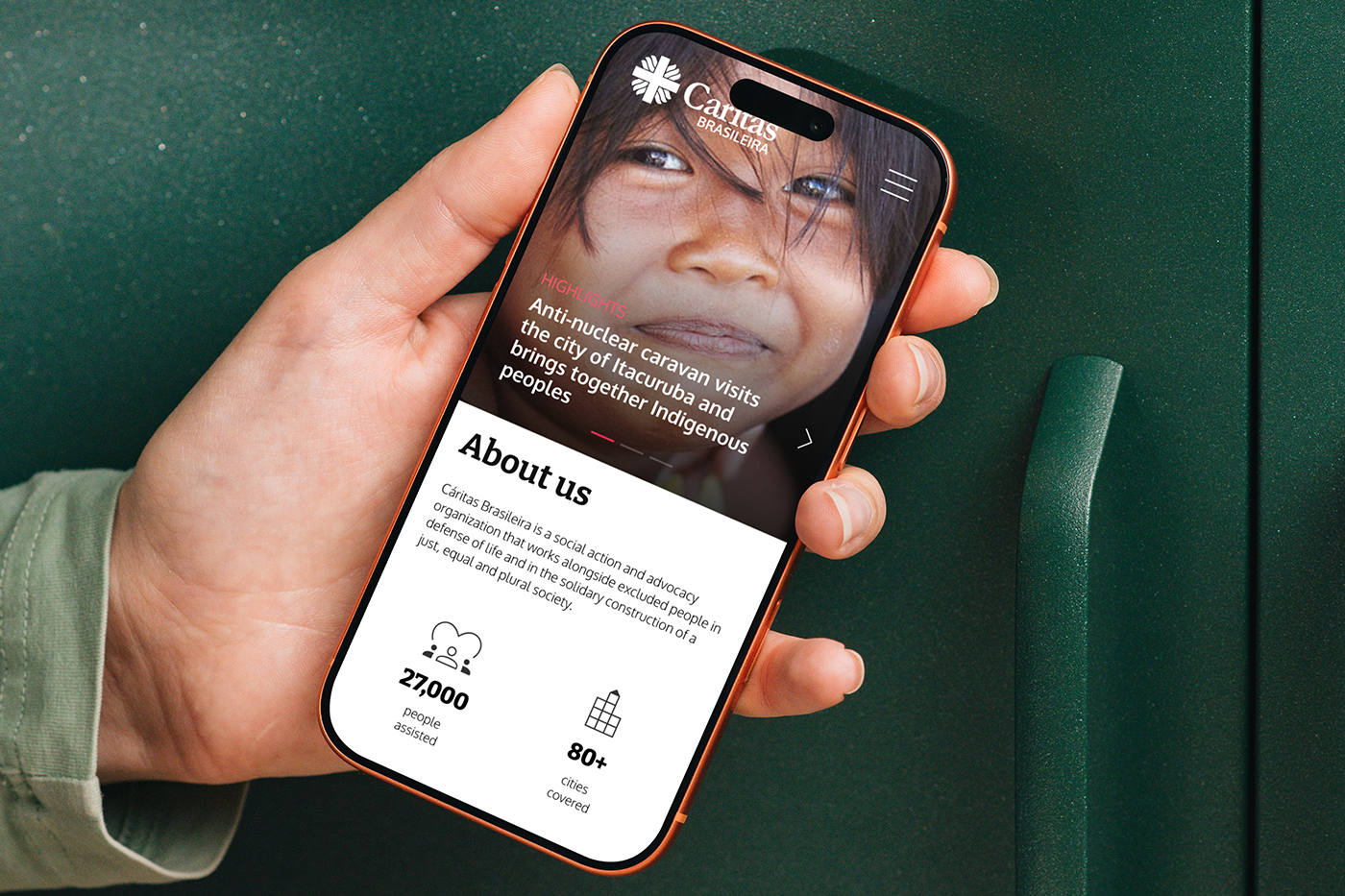 A hand holding a phone with the Caritas Brasileira mobile site open on the homepage